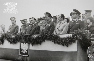 1.Tytuł tematu:  uroczystość wręczenia sztandaru Łużyckiej Brygadzie Wojsk Ochrony Pogranicza
2.Opis obrazu: uczestnicy uroczystości wręczenia sztandaru Łużyckiej BrygadzieWOP na trybunie honorowej
3.Data wydarzenia: 12.05.1963 r.
4.Osoby widoczne: dowódca WOP gen. bryg. Eugeniusz Dostojewski (trzeci od lewej),ppłk dypl. Henryk Chmielak (piąty od lewej) 
5.Hasła przedmiotowe: Wojska Ochrony Pogranicza, sztandary
6.Opis techniczny: 12 x 18 cm, oryginał, czarnobiałe
7.Autor: brak danych
8.Miejsce przechowywania/właściciel: Sala Tradycji Ośrodka Szkoleń  Specjalistycznych SG w Lubaniu