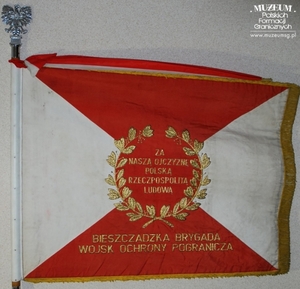 1.Nazwa własna: sztandar Bieszczadzkiej Brygady Wojsk Ochrony Pogranicza
2.Charakterystyka przedmiotu: płat wykonany z jedwabnego rypsu (130 x 100 cm), barwy biała i czerwona zostały ułożone w cztery trójkąty, na stronie głównej - pośrodku znajduje się orzeł bez korony, otoczony złotymi gałązkami wawrzynu. Na stronie odwrotnej – w centrum widnieje dewiza „Za naszą ojczyznę Polską Rzeczpospolitą Ludową”, otoczona złotym wieńcem wawrzynowym. Poniżej (na czerwonym tle) umieszczony jest napis „Bieszczadzka Brygada Wojsk Ochrony Pogranicza”
3.Hasła przedmiotowe: Wojska Ochrony Pogranicza, sztandary 
4.Czas powstania: brak danych
5.Miejsce przechowywania/właściciel: Bieszczadzki Oddział Straży Granicznej w Przemyślu